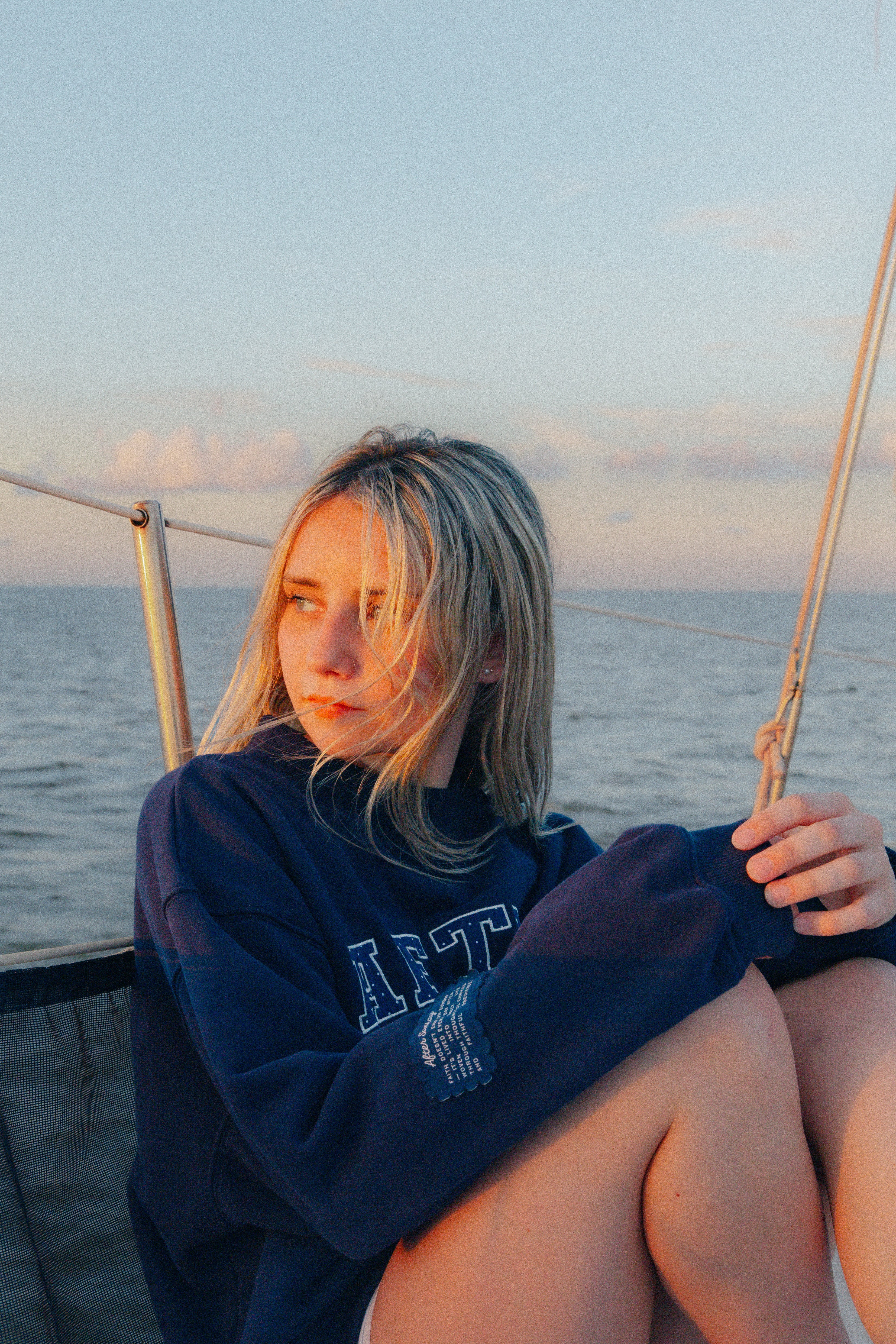 A young woman with blonde hair sitting on a boat deck, wearing an oversized navy navy sweatshirt with 'AFTER SUNDAY' printed on the front.

