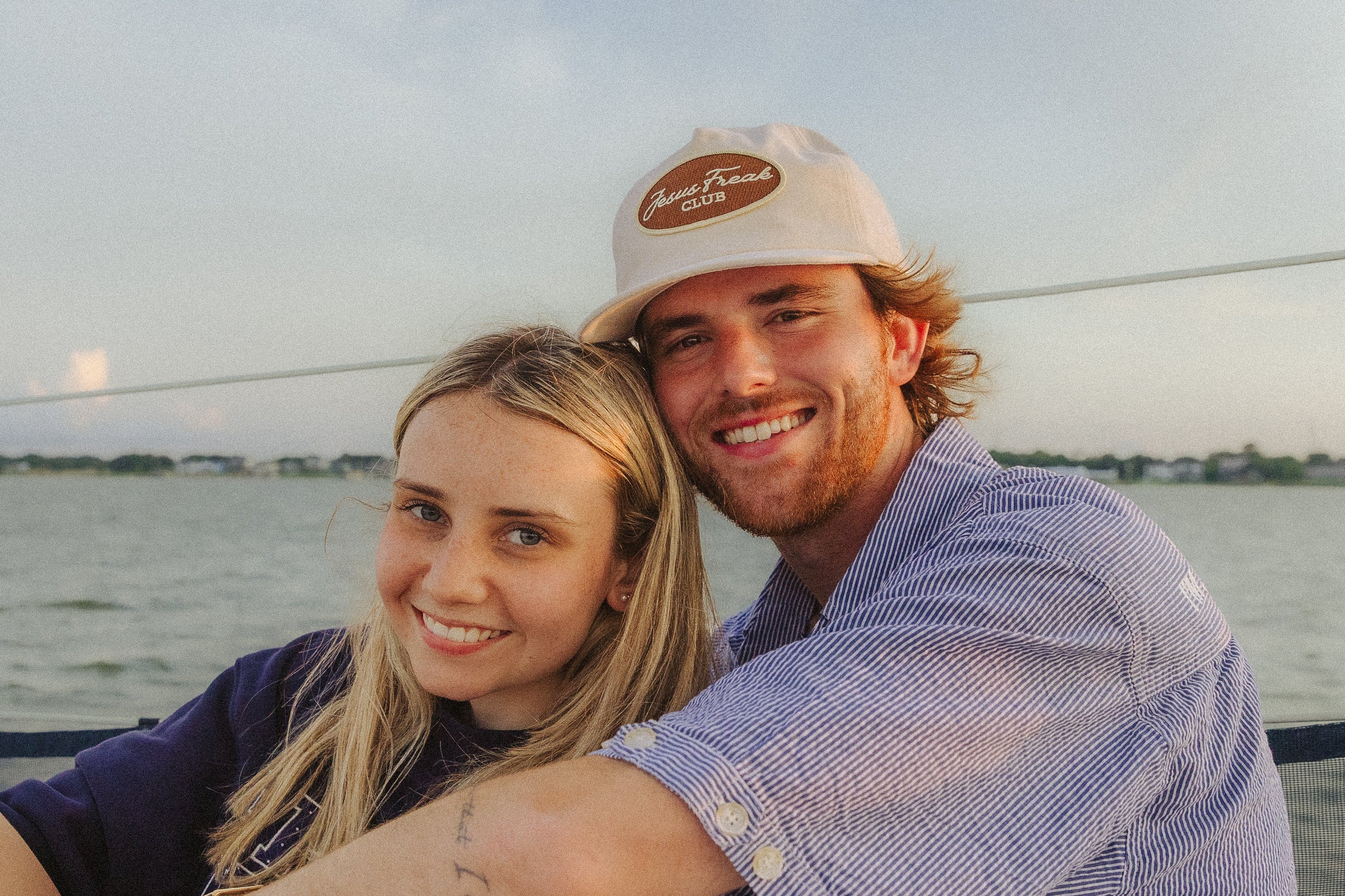 A smiling couple posing together on a boat with water and a cloudy sky in the background. The man wears a light blue button-up shirt and a beige baseball cap, while the woman wears a dark navy top.