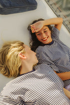 Two women lying on a white deck, wearing matching navy and white striped shirts. One has blonde hair and is looking up at the other, who has dark hair and is smiling while shielding her eyes from the sun.