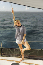 A woman posing on the deck of a sailboat, wearing a navy and white horizontally striped t-shirt and white shorts, with the blue ocean water in the background.

