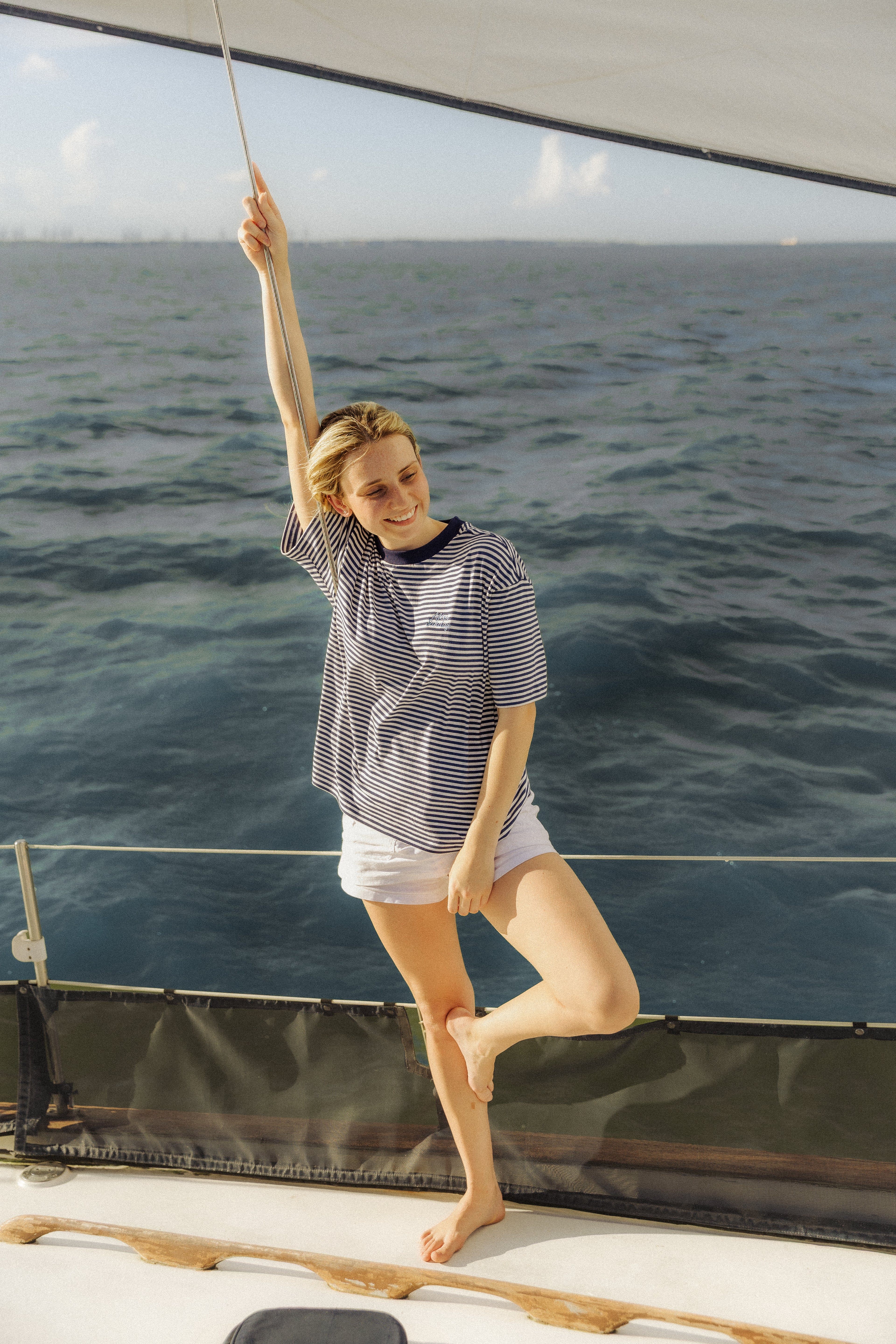 A woman posing on the deck of a sailboat, wearing a navy and white horizontally striped t-shirt and white shorts, with the blue ocean water in the background.

