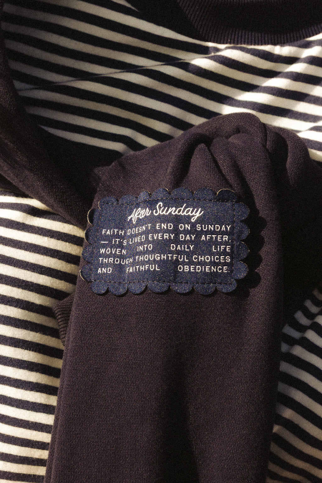 A close-up of a dark navy knit garment laid over a black-and-white striped fabric, with a sewn label reading 'After Sunday' and a scripture quote.

