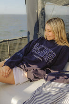 A woman sitting on a boat deck wearing a navy blue sweatshirt with 'AFTER SUNDAY' printed in white varsity lettering across the chest.

