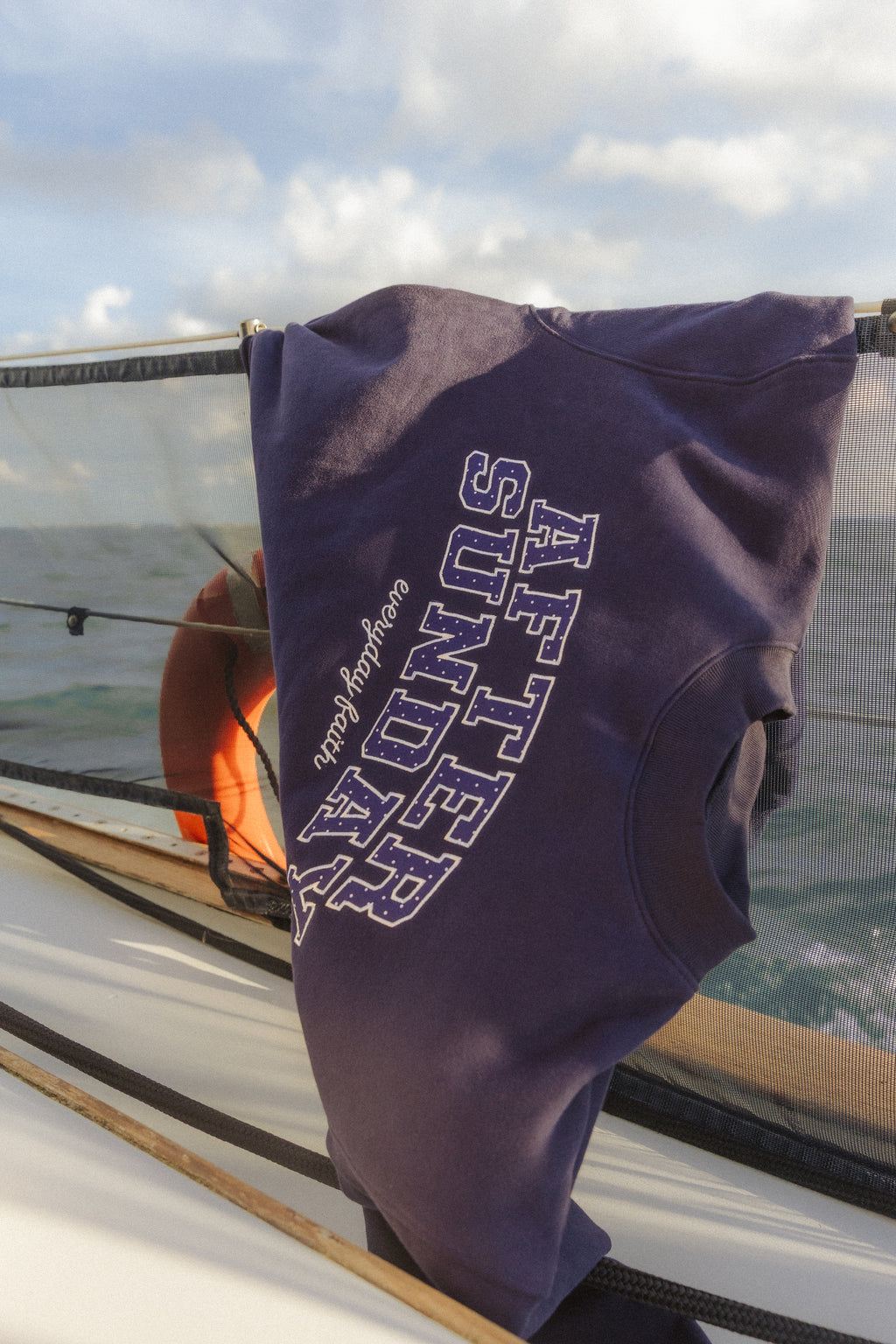 A navy blue crewneck sweatshirt with 'AFTER SUNDAY' printed in white arched block letters on the back, hanging on a boat railing with the ocean and cloudy sky in the background.

