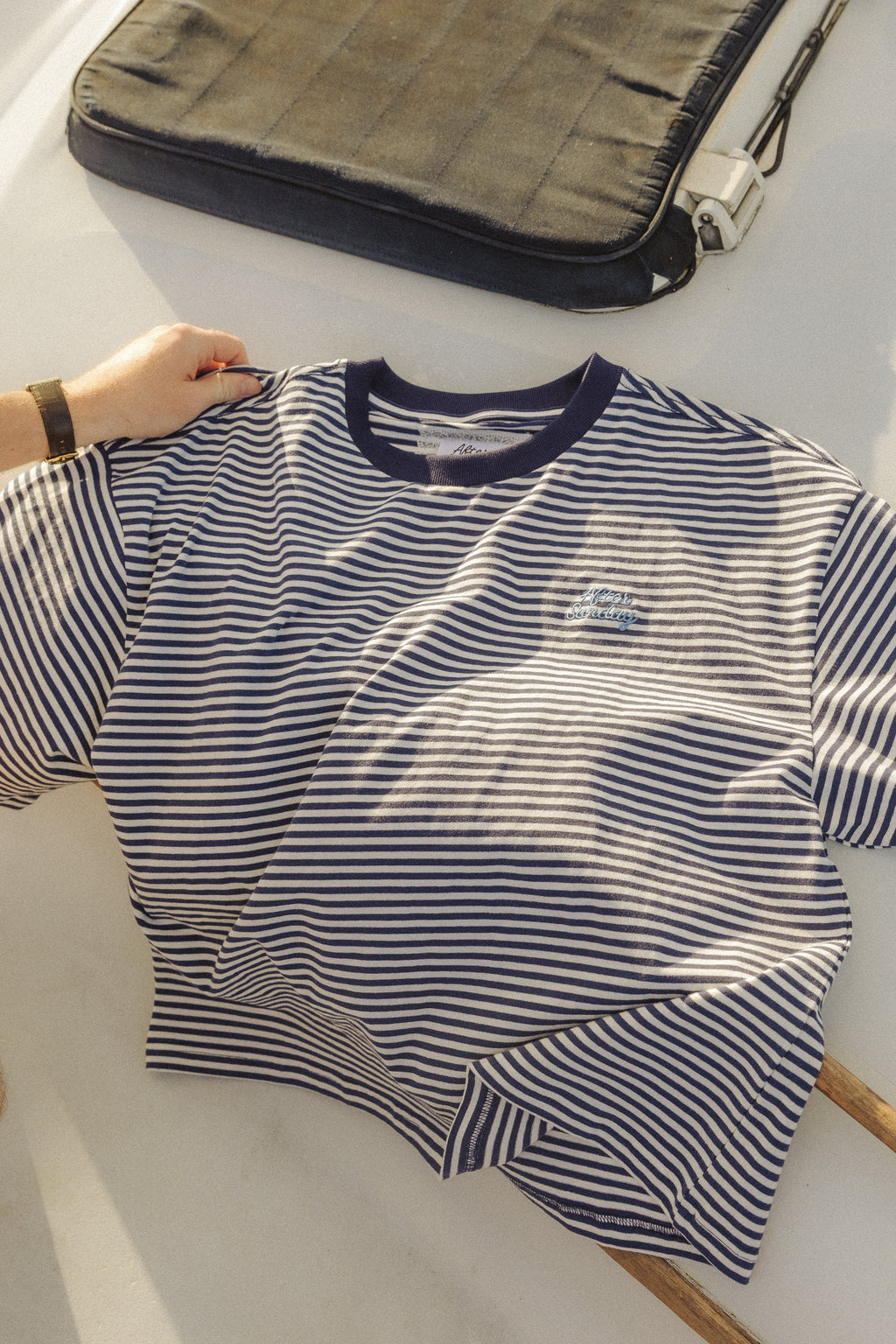 A navy and white horizontally striped short-sleeve t-shirt with a dark navy crew neck, laid flat on a light surface with a hand holding one sleeve. A dark gray laptop sleeve is visible in the background.

