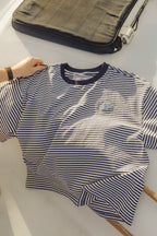 A navy and white horizontally striped short-sleeve t-shirt with a dark navy crew neck, laid flat on a light surface with a hand holding one sleeve. A dark gray laptop sleeve is visible in the background.

