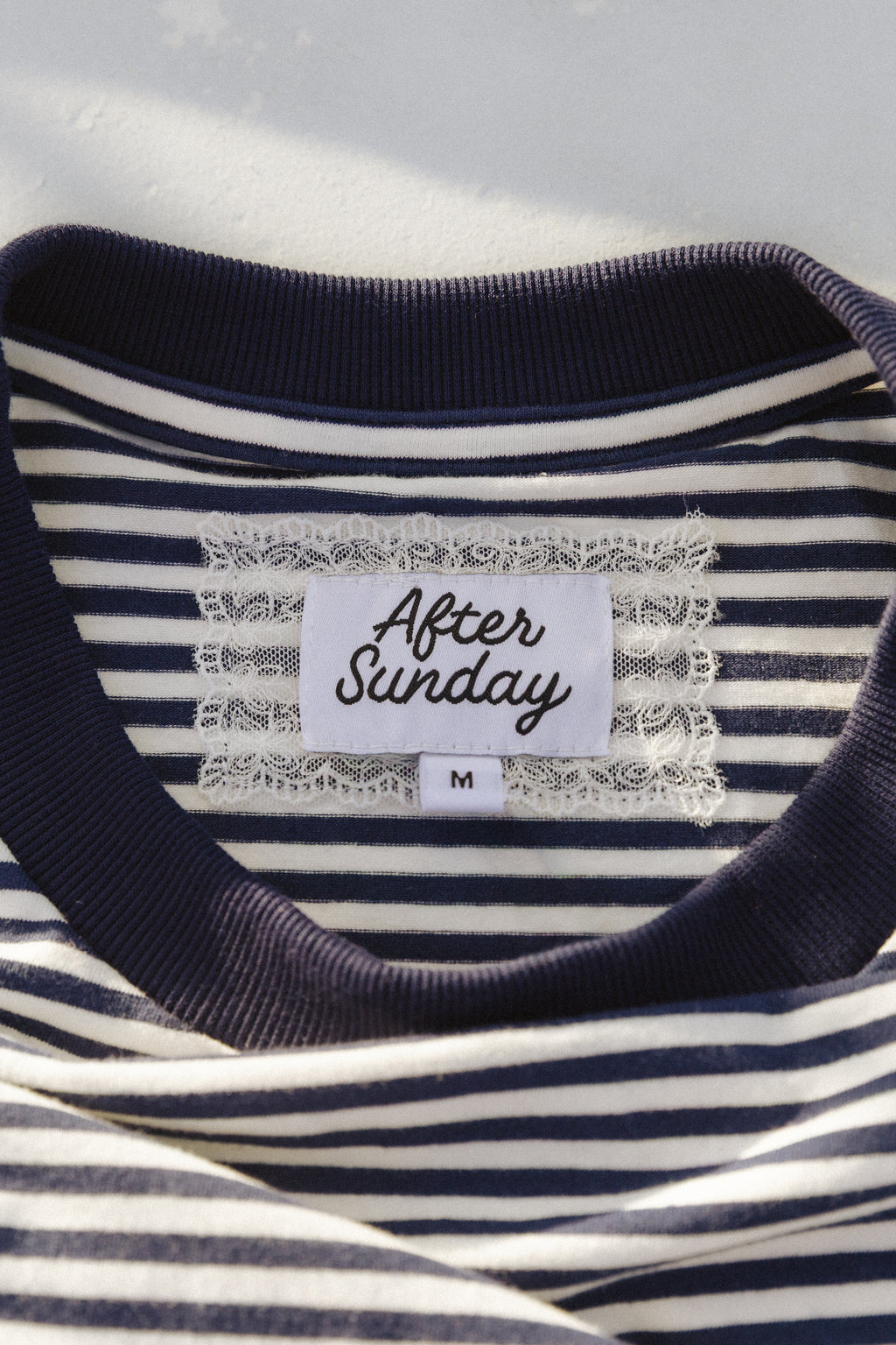 Close-up of a navy and white striped t-shirt with a white lace-trimmed neck label that reads 'After Sunday' and size 'M'.


