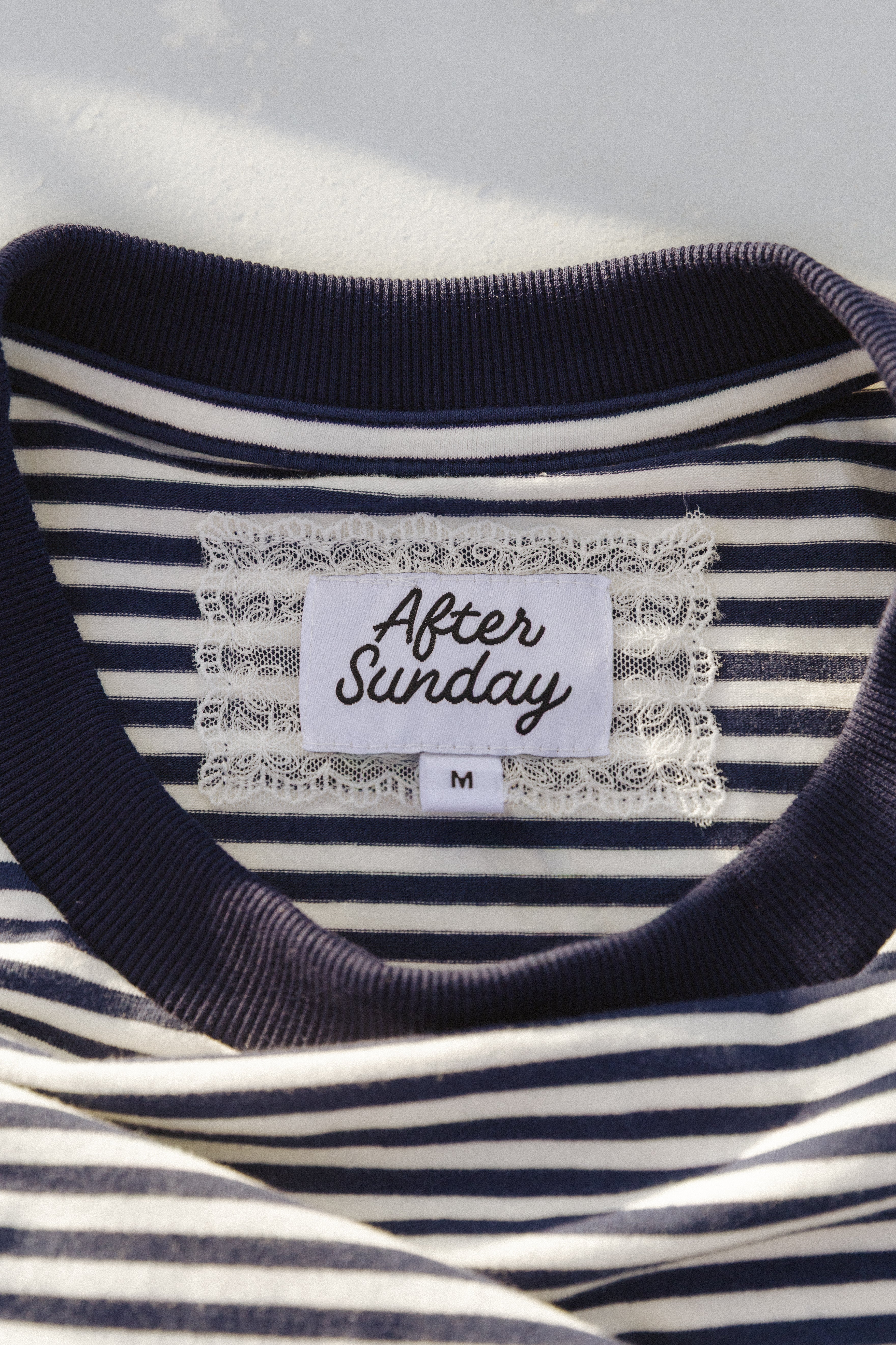 Close-up of a navy and white striped t-shirt with a white lace-trimmed neck label that reads 'After Sunday' and size 'M'.


