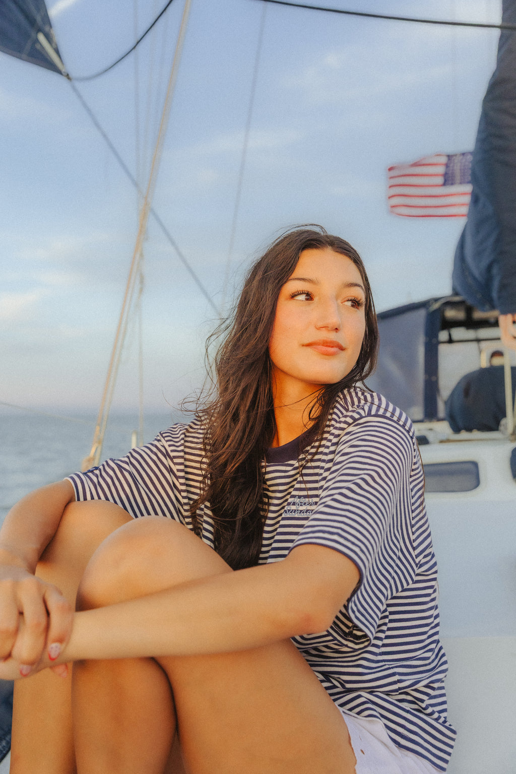 A woman sitting on the deck of a sailboat wearing a navy and white striped t-shirt and white shorts, looking off-camera against a soft sky background.

