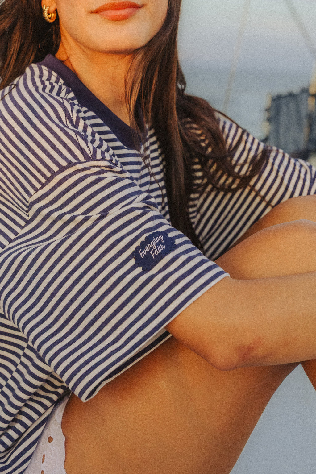 A close-up lifestyle shot of a woman wearing a navy and white striped short-sleeve t-shirt with a small embroidered 'Everyday Faith" on the sleeve, set against a soft-focus outdoor background.

