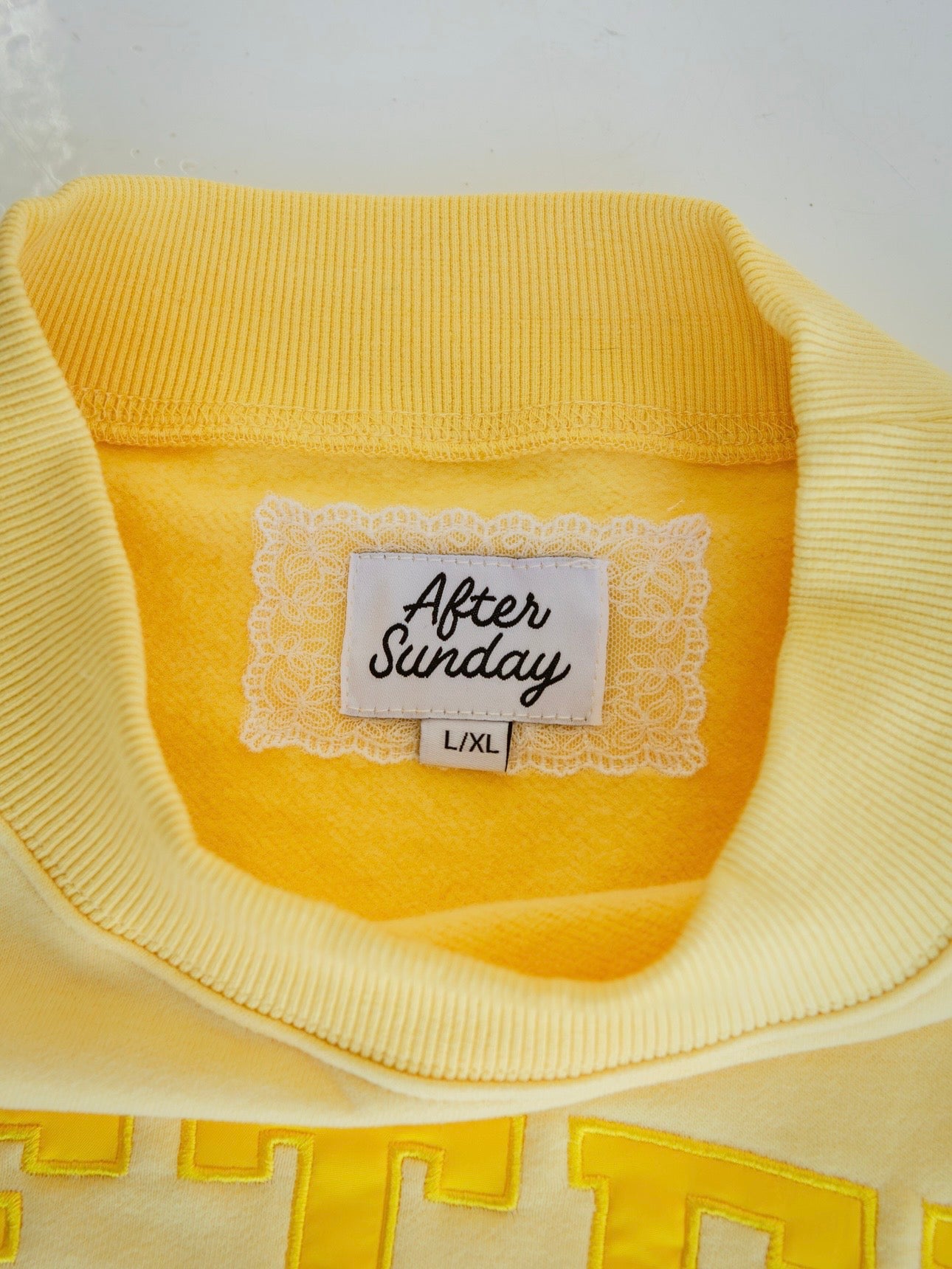 Close-up of a yellow ribbed crewneck sweatshirt showing the inner neck label with 'After Sunday' and size 'L/XL'.

