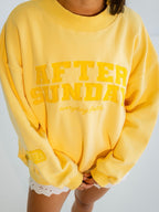 A person wearing a bright yellow oversized sweatshirt with 'AFTER SUNDAY' and 'everyday faith' printed in yellow on the front, paired with white lace hem shorts against a light gray background.