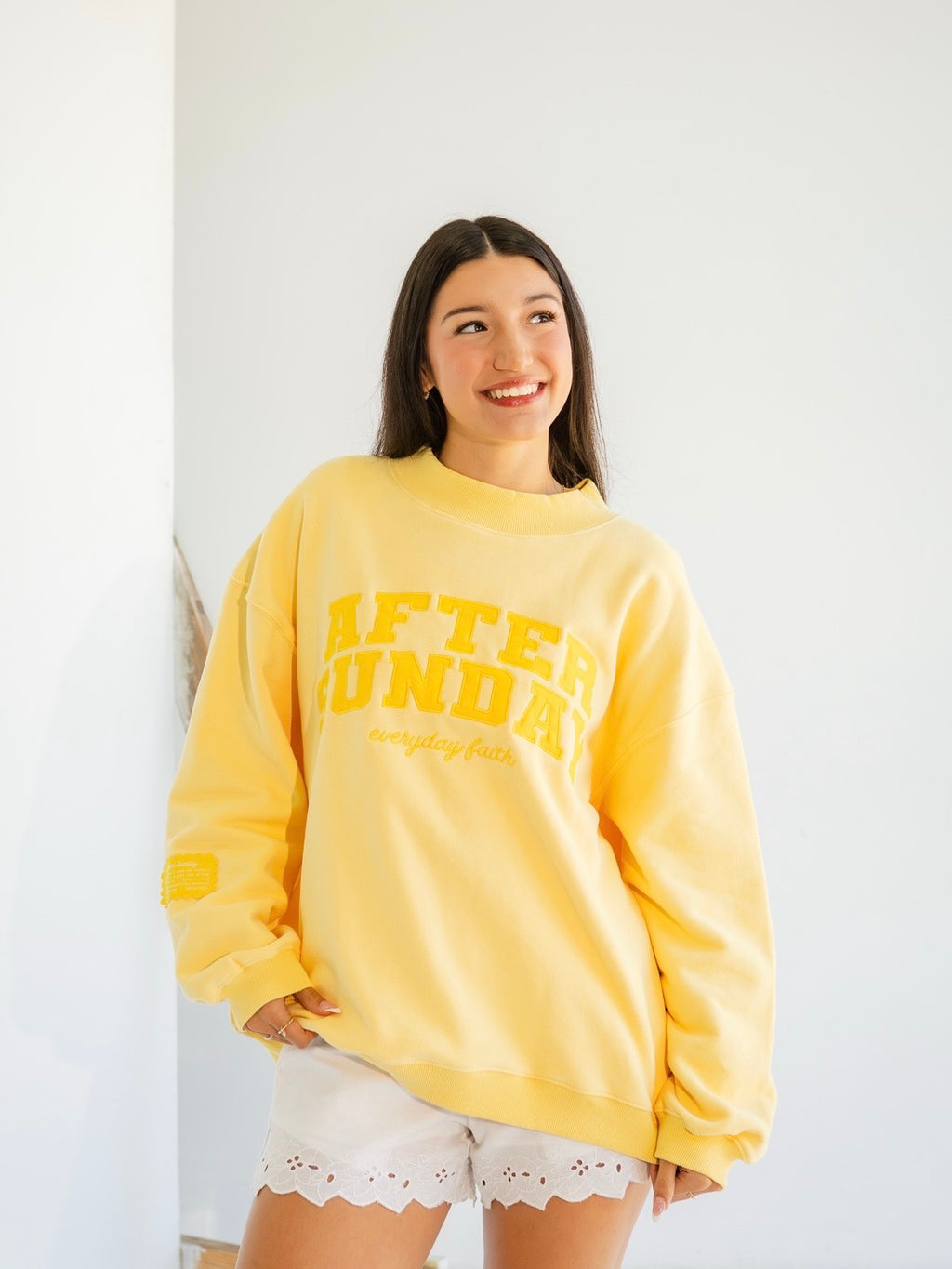 A woman wearing a bright yellow crewneck sweatshirt with 'AFTER SUNDAY' printed in yellow on the front, paired with white lace-trim shorts, standing against a plain white wall.

