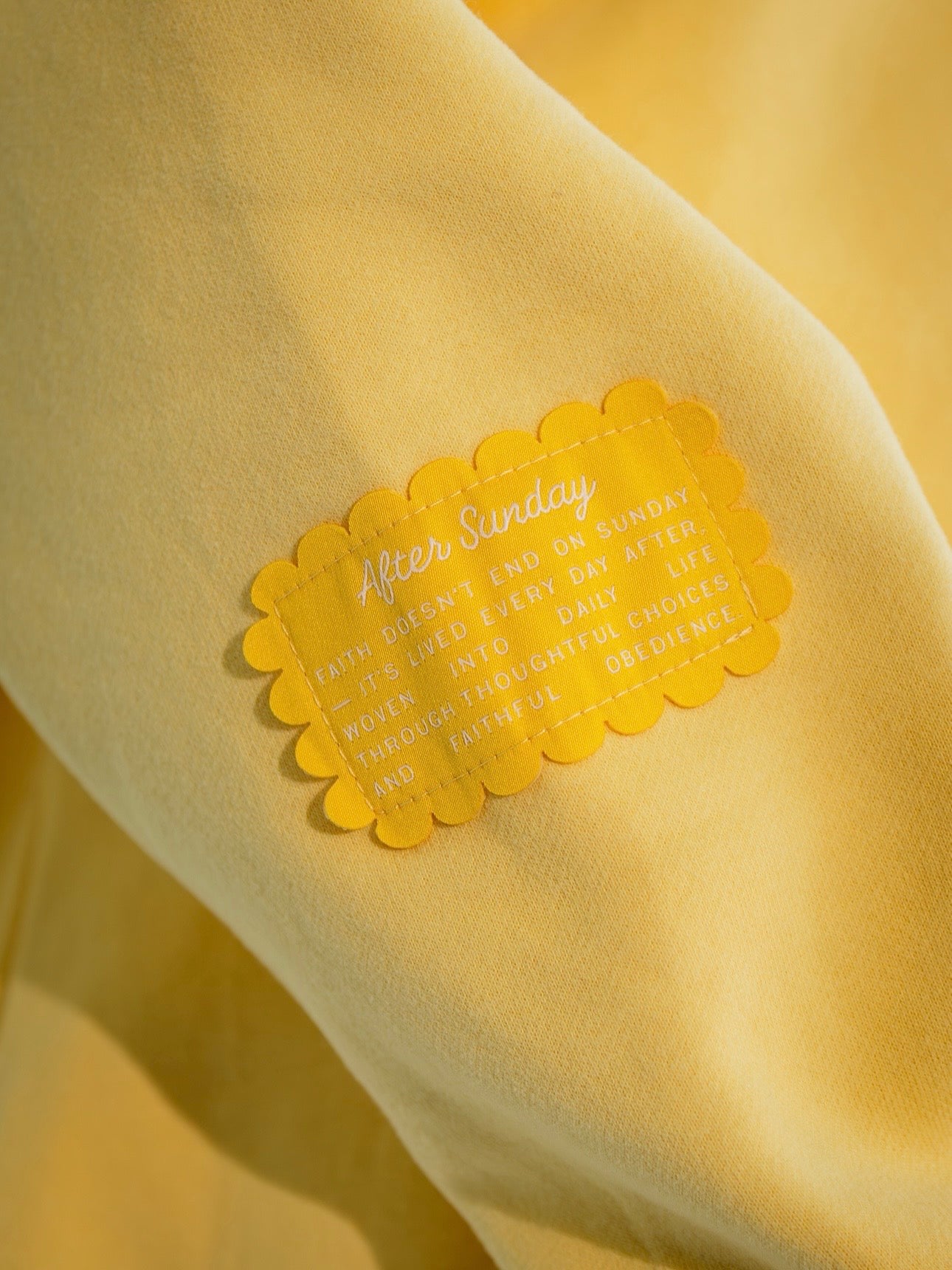 A close-up of a yellow garment with a scalloped yellow patch that reads 'After Sunday' and a motivational quote about faith, life, and obedience.


