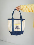 A cream canvas tote bag with navy blue handles, trim, and bottom panel, featuring a yellow gingham pocket and 'After Sunday' embroidered in navy on the front. A person's hand and arm wearing a yellow sweatshirt are holding the bag against a light gray background.