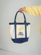 A cream canvas tote bag with navy blue handles, trim, and bottom panel, featuring a yellow gingham pocket and 'After Sunday' embroidered in navy on the front. A person's hand and arm wearing a yellow sweatshirt are holding the bag against a light gray background.