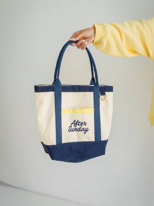 A cream canvas tote bag with navy blue handles, trim, and bottom panel, featuring a yellow gingham pocket and 'After Sunday' embroidered in navy on the front. A person's hand and arm wearing a yellow sweatshirt are holding the bag against a light gray background.