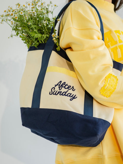 A person in a yellow sweatshirt carries a cream and navy tote bag with 'After Sunday' embroidered on the front, holding a bunch of small white flowers against a plain light background.