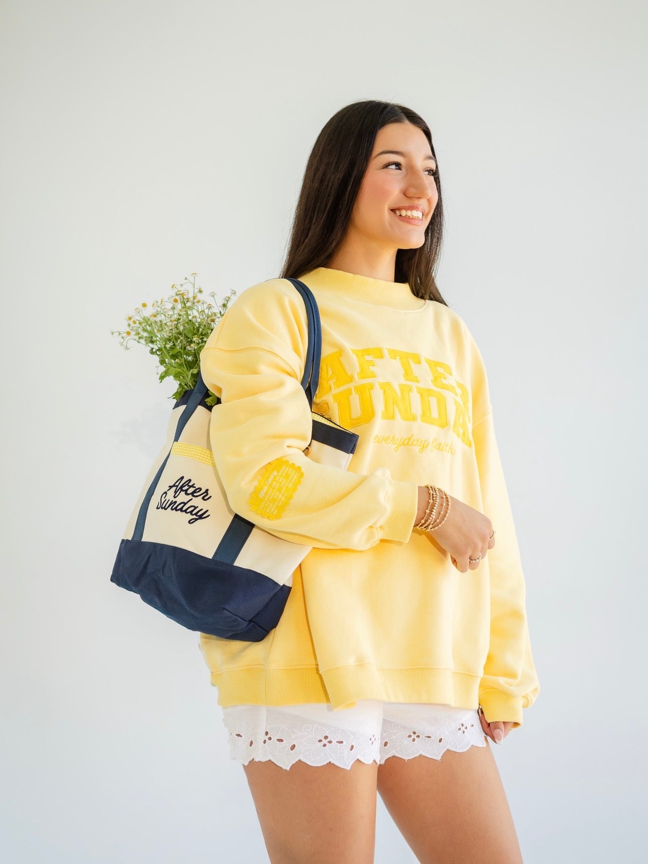 A woman wearing a yellow sweatshirt with 'AFTER SUNDAY' printed in yellow on the front, white lace-trimmed shorts, and carrying a navy and cream tote bag, posed against a light gray studio background.
