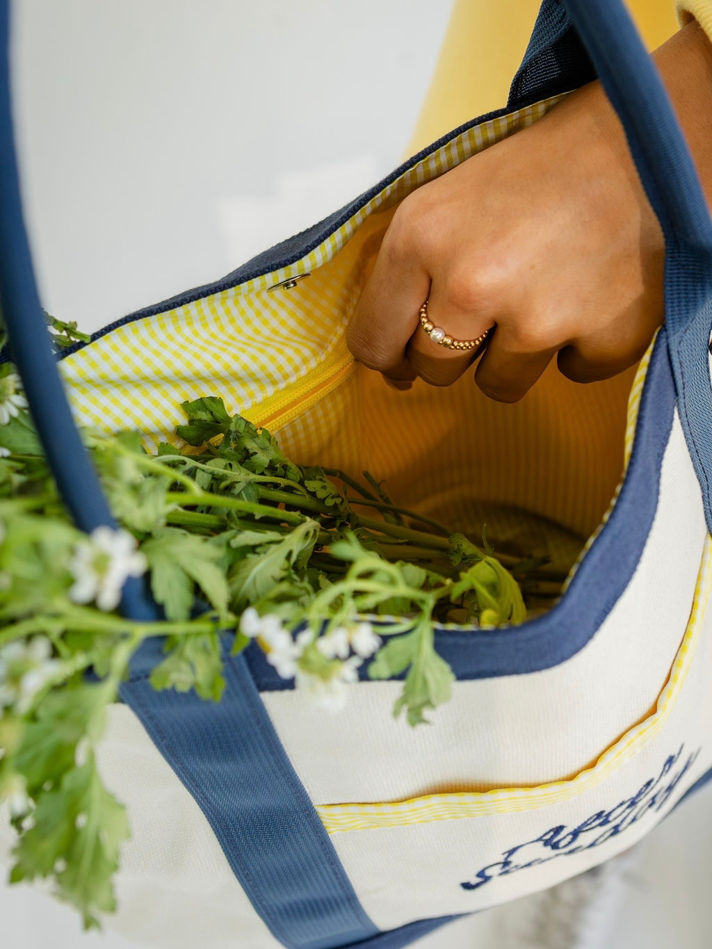 A close-up of a hand reaching into a cream canvas tote bag with navy blue straps and trim, featuring a yellow-and-white gingham interior lining and green leafy greens inside.