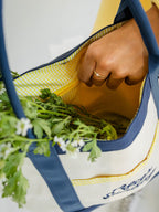 A close-up of a hand reaching into a cream canvas tote bag with navy blue straps and trim, featuring a yellow-and-white gingham interior lining and green leafy greens inside.