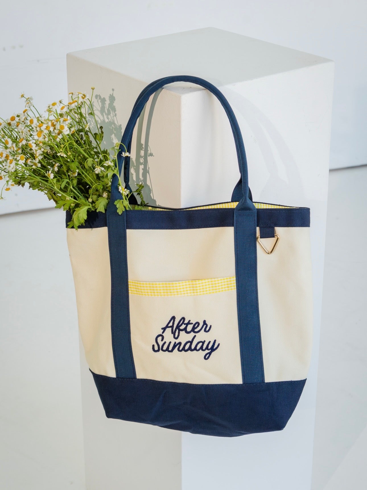 A cream canvas tote bag with navy blue trim, handles, and bottom panel, featuring a yellow-gold horizontal stripe across the front pocket and the embroidered text 'After Sunday' in navy. The bag is shown hanging from a white display block with a small bouquet of white flowers tucked into the side.