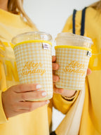 Two people in yellow sweaters hold clear plastic takeaway cups with yellow gingham sleeve covers that read 'After Sunday' in gold script.


