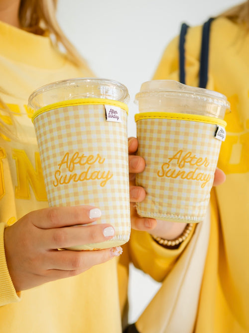 Two people in yellow sweaters hold clear plastic takeaway cups with yellow gingham sleeve covers that read 'After Sunday' in gold script.

