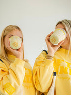 Two women in matching butter yellow sweatshirts drink from matching paper cups with yellow text that reads "Jesus Loves You"!

