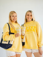 Two women standing side-by-side wearing matching yellow sweatshirts with 'After Sunday' embroidered in yellow on the front, holding yellow patterned coffee cups and wearing navy and cream tote bags against a plain white background.