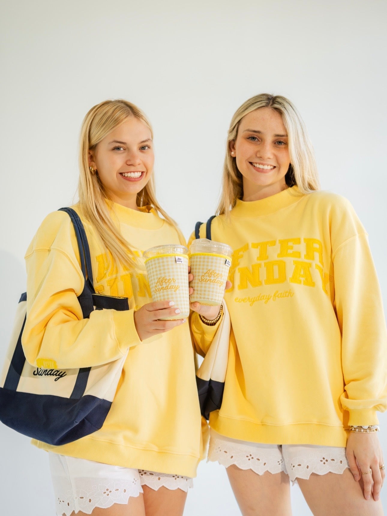 Two women standing side-by-side wearing matching yellow sweatshirts with 'After Sunday' embroidered in yellow on the front, holding yellow patterned coffee cups and wearing navy and cream tote bags against a plain white background.