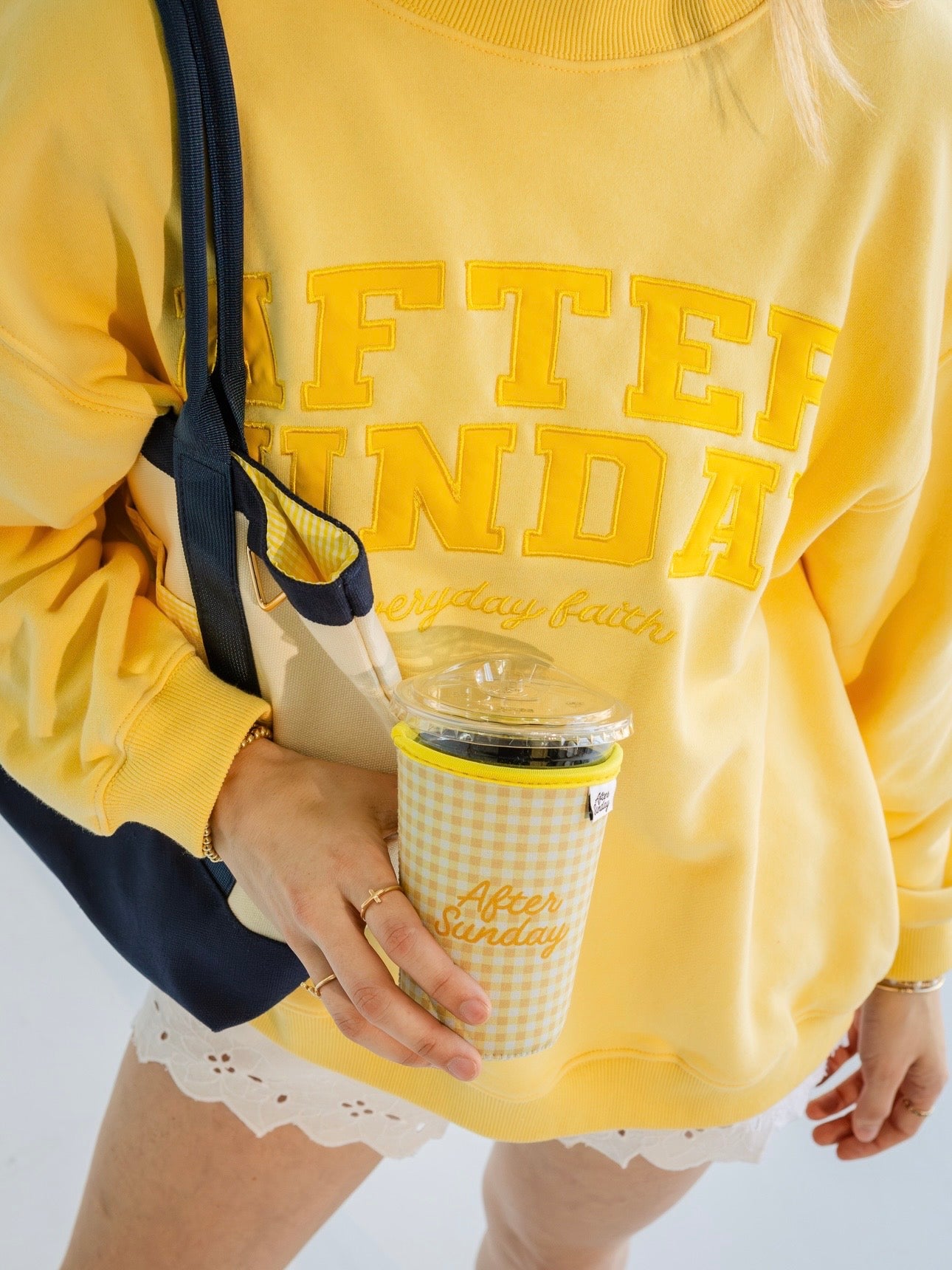 A person wearing a bright yellow sweatshirt with large yellow text reading 'AFTER SUNDAY' and smaller script text below, holding a gingham coffee cup while carrying a navy tote bag.