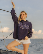 A woman posing on a boat wearing a dark navy sweatshirt with white polkadot graphic lettering that reads 'After Sunday' and white shorts, with the ocean and sky in the background.

